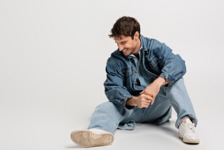 full length of stylish man in denim jacket and jeans sitting and laughing on grey,stock image